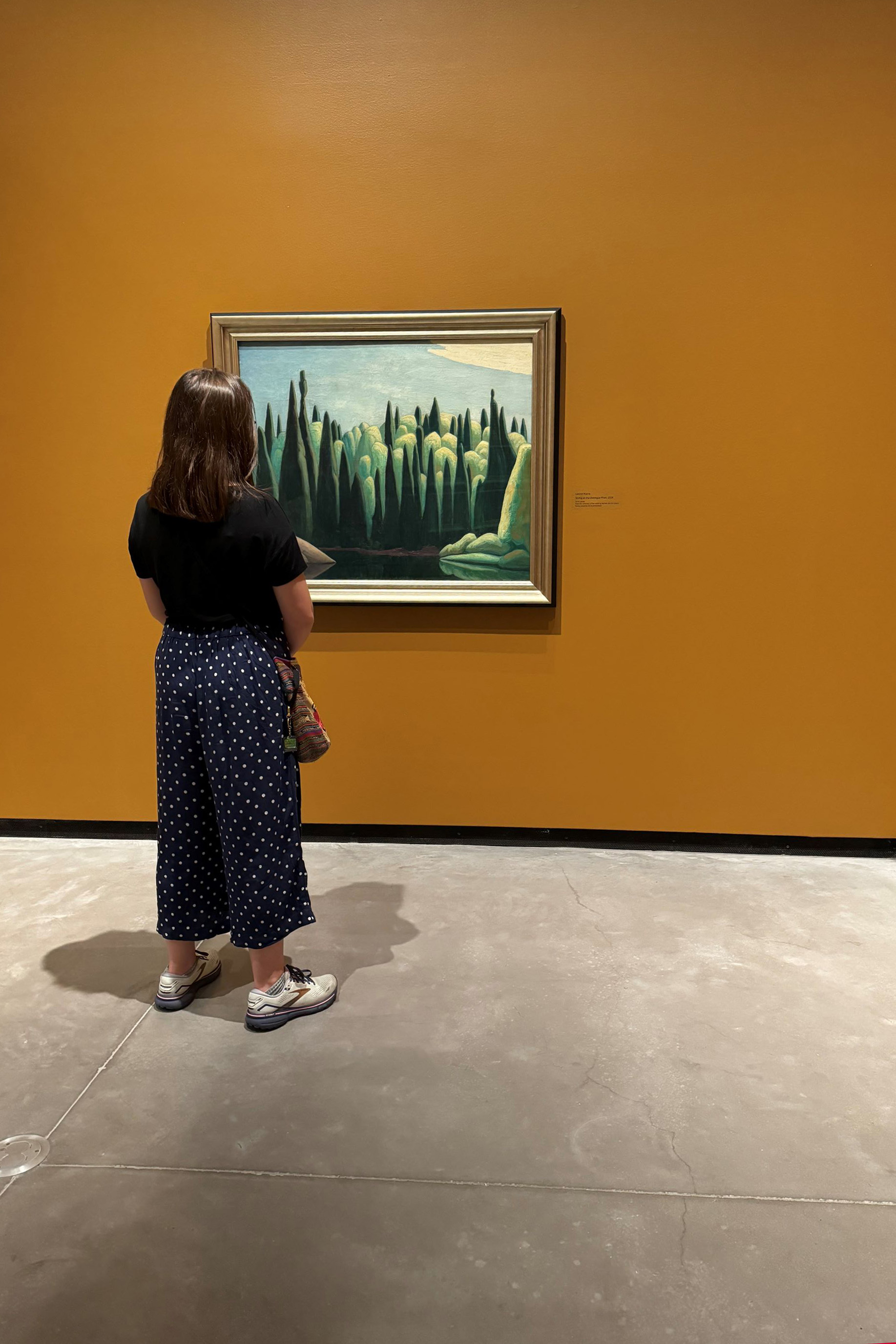 A woman with black shirt and blue polkadot pants looks at a painting  of green trees and a blue sky hanging on an orange wall