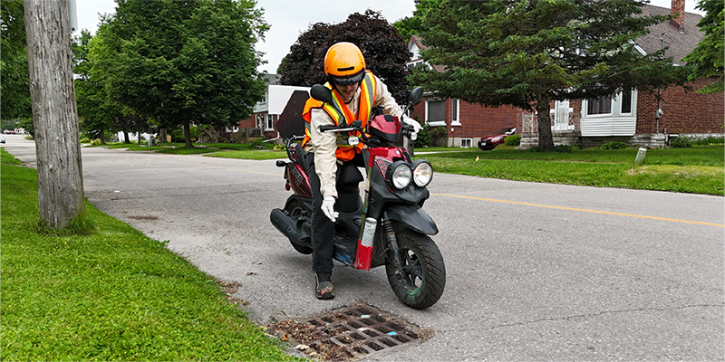 Man on a motorized scooter drops larvicide into roadside catchbasin