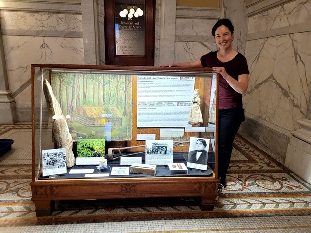 Photo of a white woman with brown hair standing next to a brown exhibit case filled with artifacts and information on 175 years of Lambton County.